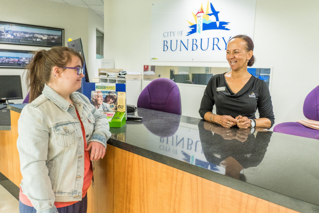 Young woman standing at counter of the administration building looking at staff member who is smiling back. City of Bunbury logo behind them as well as an office chair and computer screen.