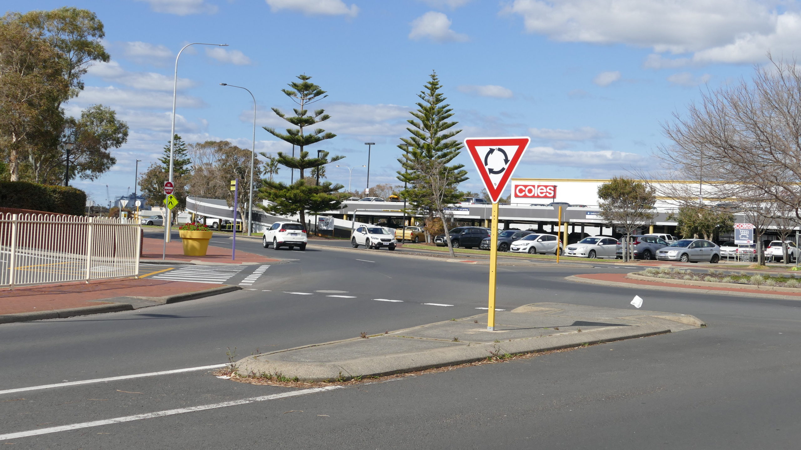 Entrance into Bunbury CBD. A road with median strip and give way sign and half a roundabout behind it. Two white cars driving along the road - one approaching the roundabout the other driving away from it. Shopping centre car park with cars in the background.