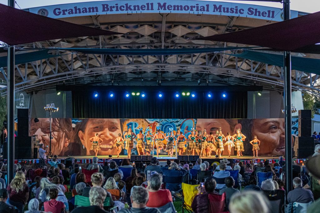 Looking across a seated crowd towards the Graham Bricknell Memorial Music Shell. Multicultural dancers are on the stage performing. The backdrop is a mural with the word 'belong' and the faces of four local women from Bunbury's multicultural community. It's later in the afternoon and the stage is lit up in lights.