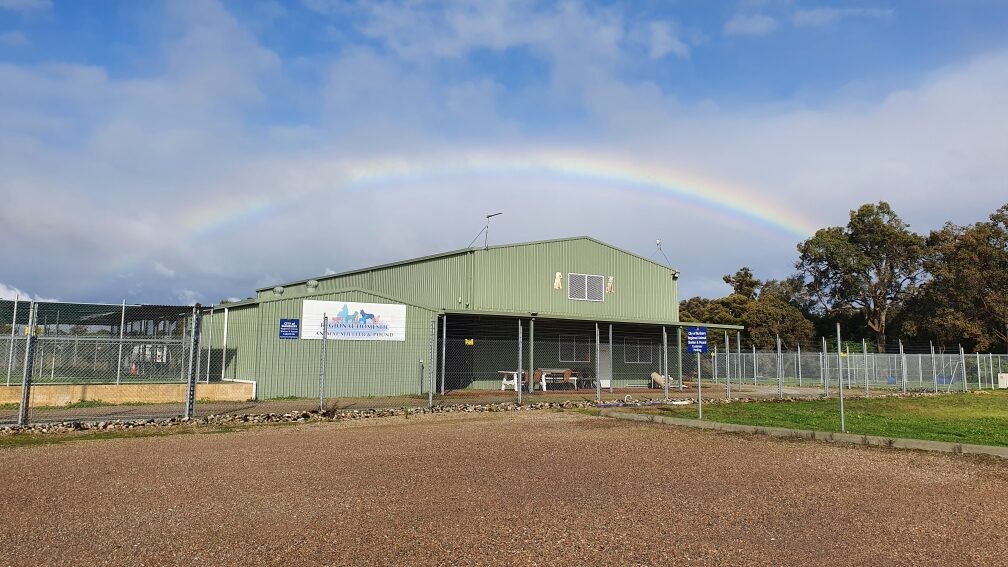 Green colorbond building with wire fencing surrounding it. Gravel driveway. Trees on the right hand side of the building and a rainbow above the building with clouds and blue sky.