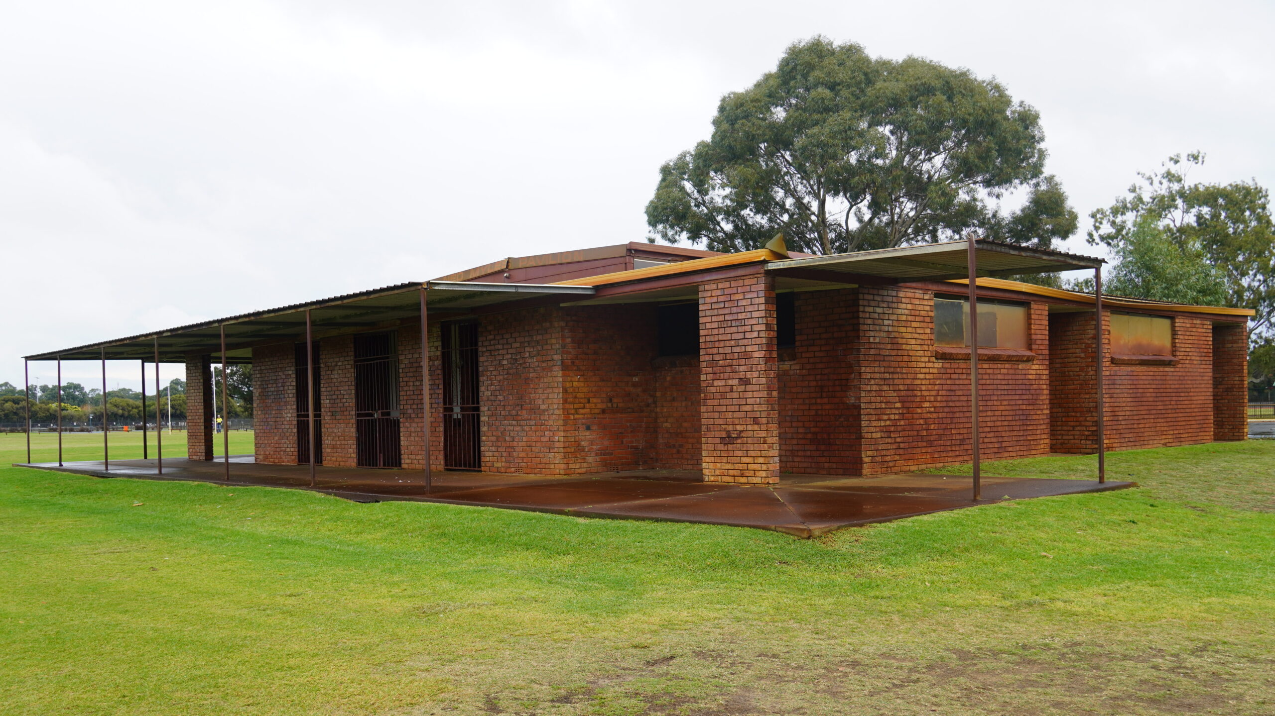 Brown brick building rusted roof and browned concrete patio located on green grass with a tree behind it