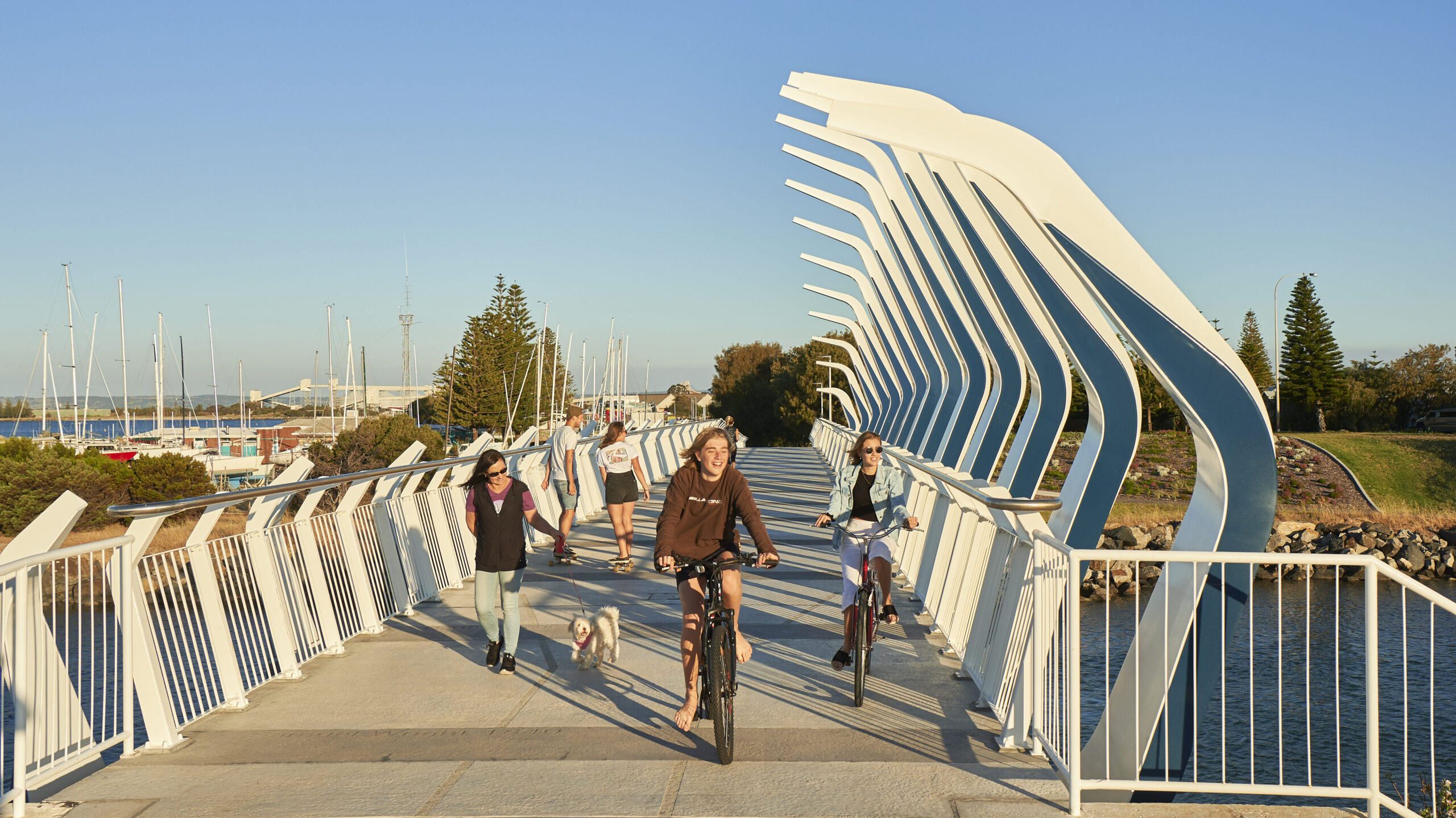 Two teenagers riding bikes along the Koombana footbridge and a woman walking a small fluffy white dog. A young man and woman in the background walking in the opposite direction. Blue sky.