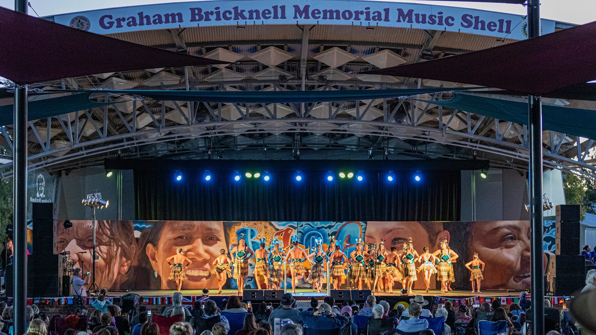 Looking across a seated crowd towards the Graham Bricknell Memorial Music Shell. Multicultural dancers are on the stage performing. The backdrop is a mural with the word 'belong' and the faces of four local women from Bunbury's multicultural community. It's later in the afternoon and the stage is lit up in lights.