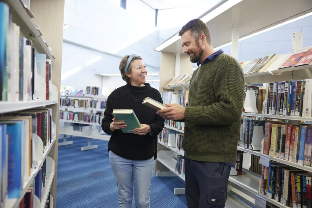 two people smiling and looking at books in library shelves filled with books