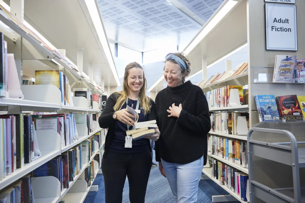 two women in library shelves looking at the same book and smiling