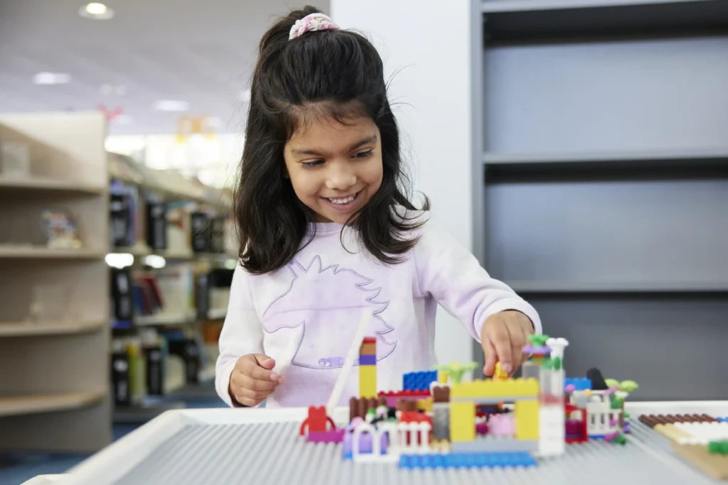 young girl building lego house with bookshelves in background