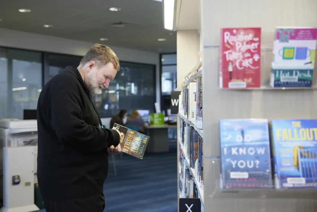man in black jacket looking at book next to shelves of books