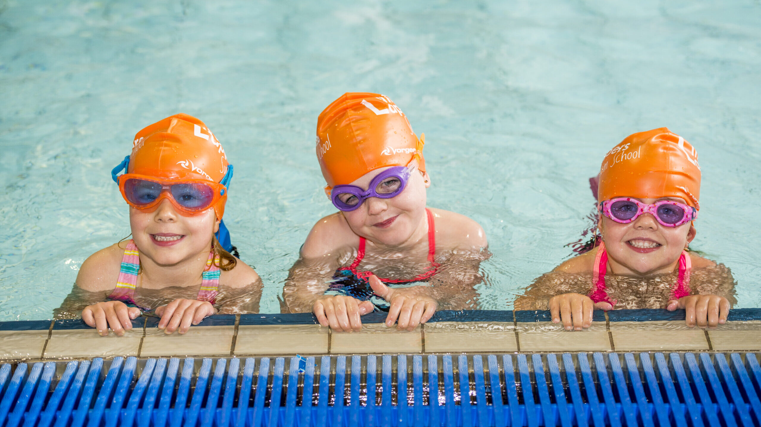 three children in orange swim caps on side of pool smiling